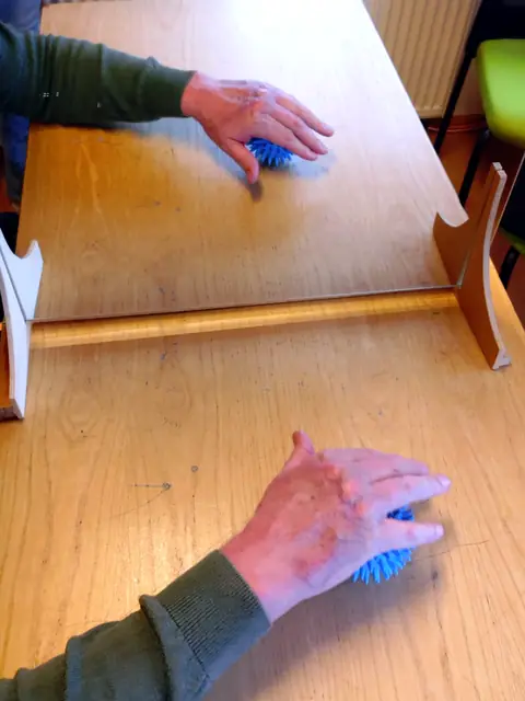 Spiegeltherapie mit Igelball: Die flache Hand rollt über einen blauen Noppenball vor dem Spiegel