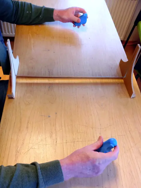 Spiegeltherapie am Tisch: Eine Hand hält Therapieknete vor einem Spiegel, oben ist ihr Spiegelbild zu sehen